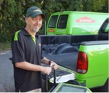 A photo of a smiling male employee wearing a black SERVPRO shirt standing in front of green SERVPRO vehicles