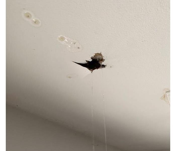 A photo of a white living room ceiling with a gaping hole and other cracks with streams of water coming out onto the floor.