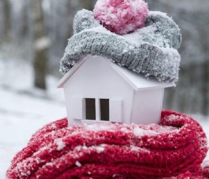 A photo of a white birdhouse sitting in the snow wrapped with a red scarf and a gray hat