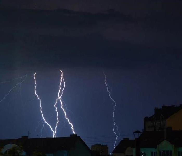 Several white lightning strikes against a dark purple sky hitting behind a group of homes.