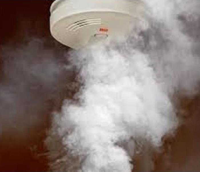 A white smoke detector on a brown wall with a large plume of smoke heading towards it.