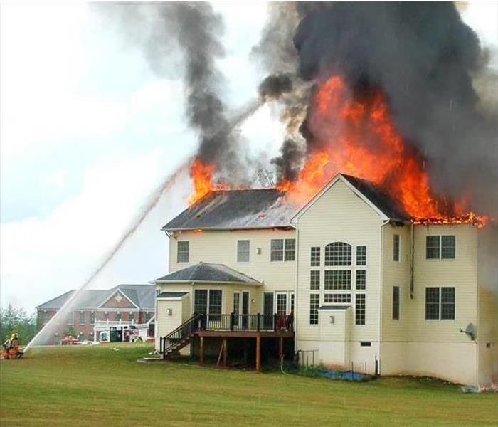 A large home with it's roof on fire while firefighters spray water on it.