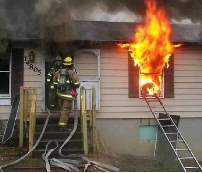 2 firemen going into a home with flames shooting out of a front window