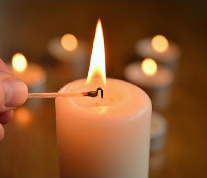 An image of a hand holding a lit match over a white candle with smaller lit candles in the background.